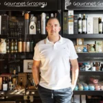 owner of gourmet groceries in front of a selection of olive oil bottles and other gourmet products
