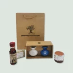 A brown paper gift bag with the logo and text "gourmetgroceries.com" sits behind a selection of gourmet products. These include a small bottle of Commandaria red wine with a red cap, a bar of soap in green and white packaging, a box containing a white ceramic bottle of extra virgin olive oil and a blue ceramic bottle of balsamic vinegar, and a jar of pine honey.