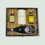 A wooden gift box containing gourmet items arranged neatly in wood wool. The items include a bottle of balsamic vinegar, a tin of chocolate, a bottle of olive oil, and a bottle of red wine.