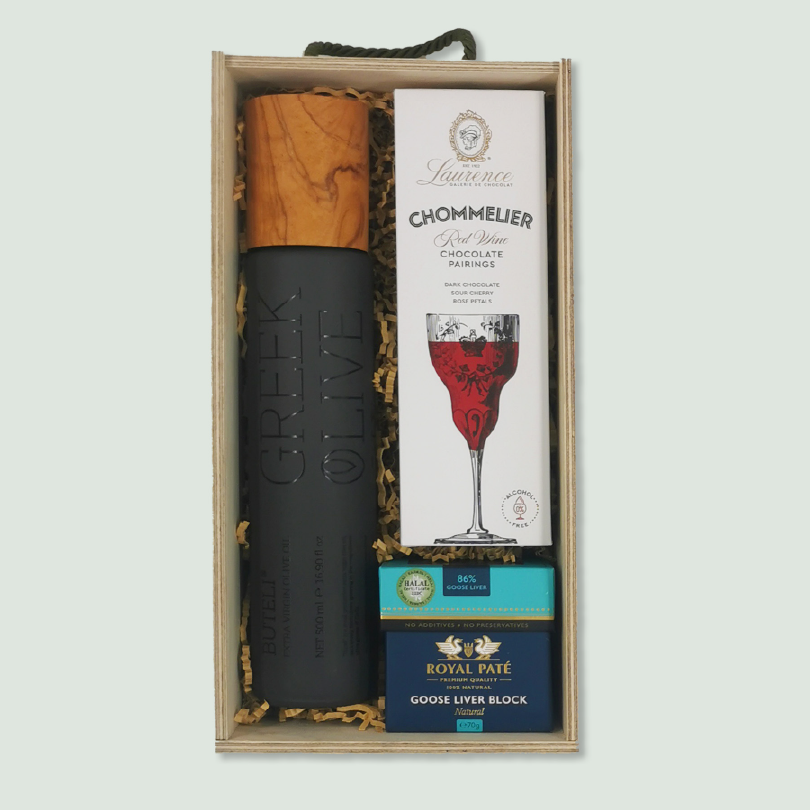 A gift box containing a bottle of Greek olive oil, a box of chocolate pairings, and a package of goose liver pâté. The items are nestled in wood wool within a wooden box with a black rope handle.