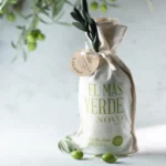 A bottle of El Más Verde Novo extra virgin olive oil positioned next to a bowl of fresh olives, emphasizing the oil's quality and origin.