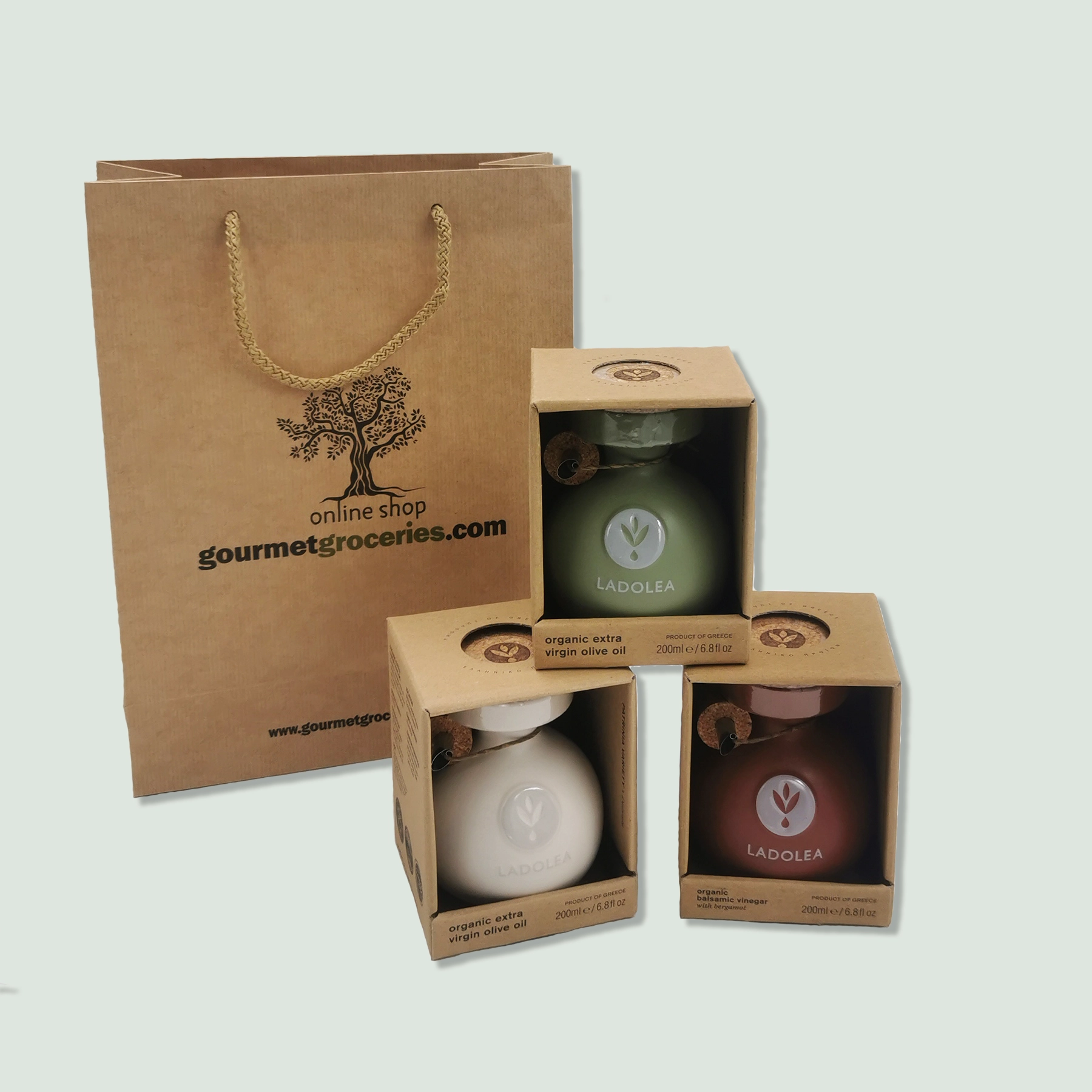 Three boxed ceramic bottles of Ladolea organic products displayed in front of a brown paper gift bag with a rope handle. The bag is branded with the logo "gourmetgroceries.com." The bottles include organic extra virgin olive oil in white and green bottles, and organic balsamic vinegar with bergamot in a red bottle.
