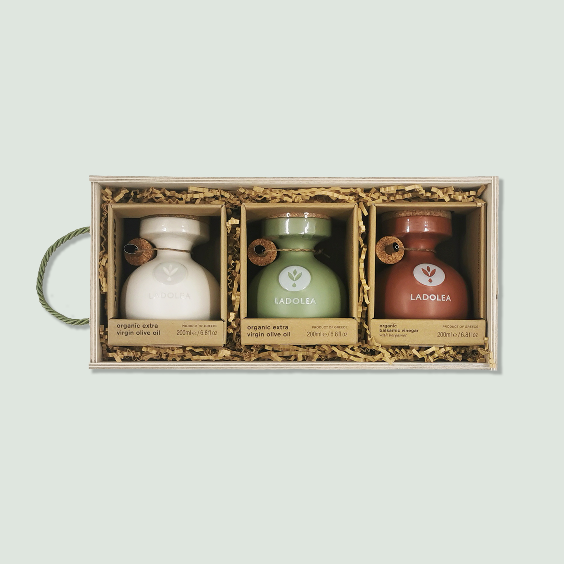 A wooden gift box containing three ceramic bottles of Ladolea organic products. The bottles are arranged side by side in wood wool. From left to right, the bottles are: organic extra virgin olive oil in a white bottle, organic extra virgin olive oil in a green bottle, and organic balsamic vinegar with bergamot in a red bottle.